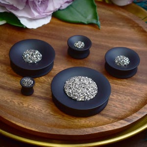 May include: A collection of black ear plugs with a silver, glittery center. The plugs are displayed on a wooden tray. The plugs vary in size, with the largest measuring approximately 1 inch in diameter.