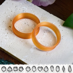 Puede incluir: Dos anillos de artesanía circulares de color naranja se muestran sobre una superficie clara y texturizada. Debajo, se ve una fila de pequeños cortadores de artesanía negros con formas geométricas, incluidos círculos, triángulos, cuadrados y corazones. Los anillos miden unos 5 cm de diámetro.