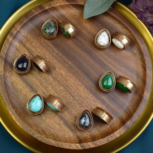 May include: A wooden tray containing eight pairs of teardrop-shaped stone and metal ear gauges. The stones are in various colours, including green, blue, black, and white.