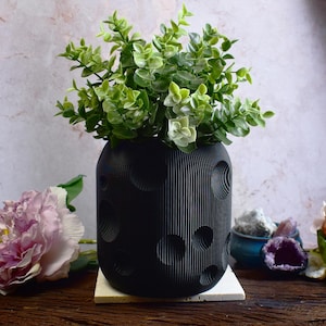 Puede incluir: Un macetero de cerámica negro con una textura lunar, con una pequeña planta verde. El macetero está sobre una superficie blanca, con flores rosas y cristales en el fondo.