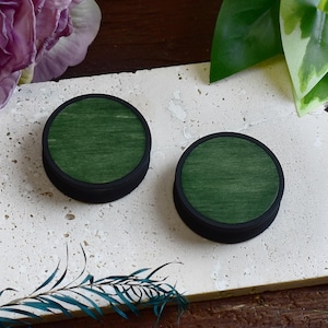 Può includere: Due tappi rotondi in legno verde scuro con bordi neri. Sono presentati su una superficie chiara e strutturata, con verde e fiori viola visibili. Questi tappi sono probabilmente per piercing all'orecchio.