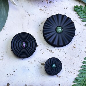 Könnte beinhalten: Drei schwarze dekorative Knöpfe mit irisierenden Zentren. Die Knöpfe haben unterschiedliche Designs: konzentrische Kreise, eine plissierte Fächerform und eine Spirale. Die Knöpfe sind auf einer weißen, strukturierten Oberfläche angeordnet.