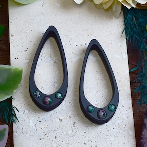 Puede incluir: Un par de pendientes en forma de lágrima de color gris oscuro. Cada pendiente presenta tres pequeñas piedras redondas, verdes y rosas moteadas. Los pendientes se muestran sobre una superficie clara y texturizada.