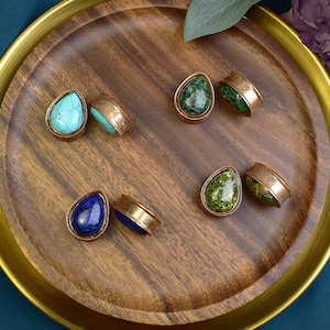 May include: Six pairs of teardrop-shaped gemstone earrings with gold-toned metal settings. The gemstones are turquoise, green, and blue.