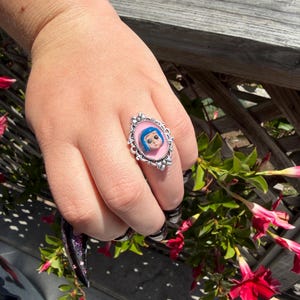 May include: Silver ring with a cartoon character's face on a pink backdrop. The ring has an ornate, oval setting with a detailed border. The ring is worn on a hand with long, black and purple nail polish.