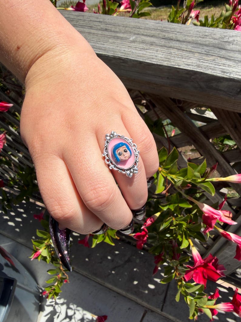 May include: Silver ring with a cartoon character's face on a pink backdrop. The ring has an ornate, oval setting with a detailed border. The ring is worn on a hand with long, black and purple nail polish.