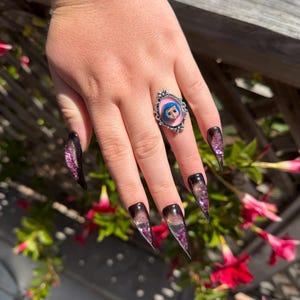 May include: A silver ring with a pink background and a cartoon character image. The nails are long, pointed, and feature a gradient design with black, purple, and clear sections. The nails have a glittery effect.