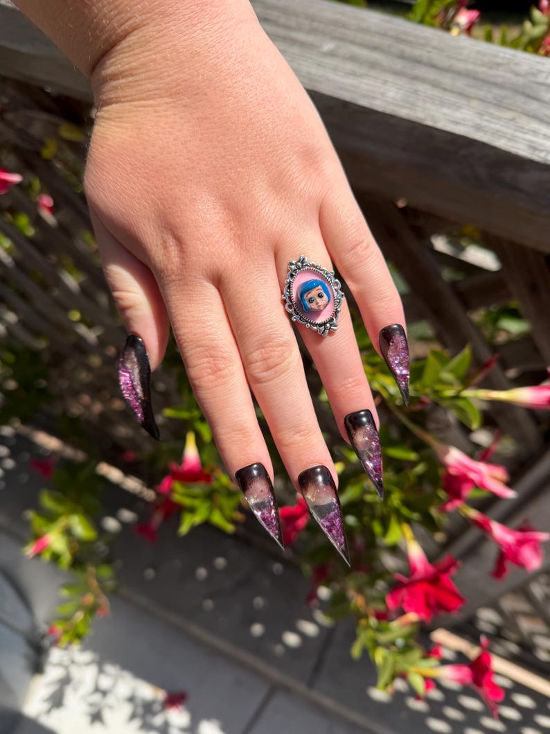 May include: A silver ring with a pink background and a cartoon character image. The nails are long, pointed, and feature a gradient design with black, purple, and clear sections. The nails have a glittery effect.