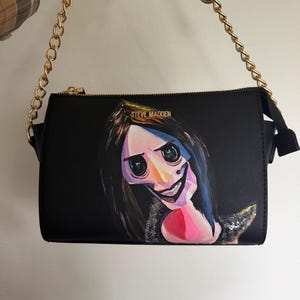 May include: Black crossbody bag with a gold chain strap and a zipper closure. The bag features a painted image of a character with large black eyes, a wide smile, and dark hair. The brand name "STEVE MADDEN" is visible on the top.