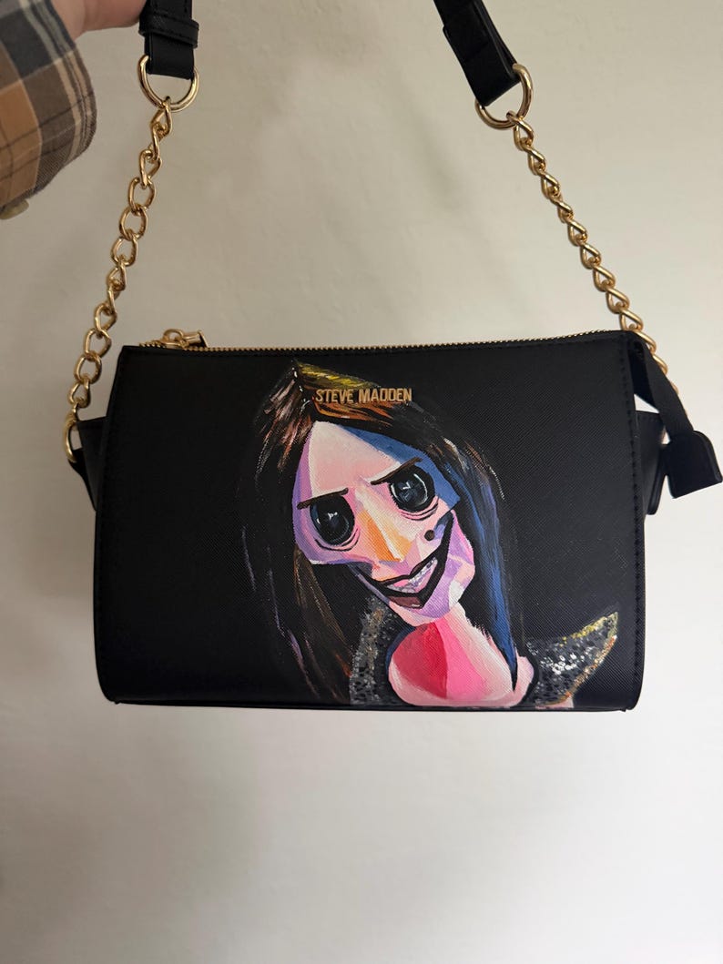 May include: Black crossbody bag with a gold chain strap and a zipper closure. The bag features a painted image of a character with large black eyes, a wide smile, and dark hair. The brand name "STEVE MADDEN" is visible on the top.