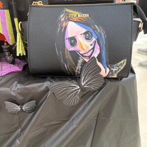 May include: Black crossbody bag with a painted illustration of a character from the movie Coraline. The bag has a gold zipper and the words "STEVE MADDEN" are printed on the front. A black and white butterfly decoration is on the table.
