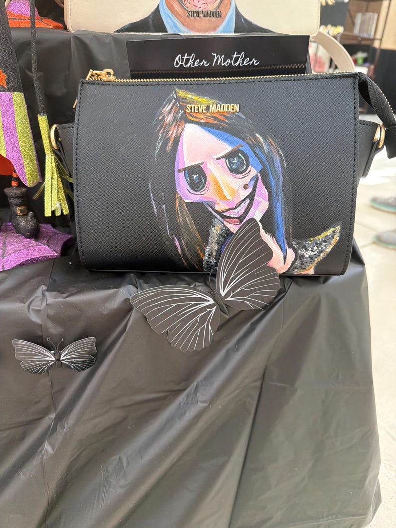 May include: Black crossbody bag with a painted illustration of a character from the movie Coraline. The bag has a gold zipper and the words "STEVE MADDEN" are printed on the front. A black and white butterfly decoration is on the table.