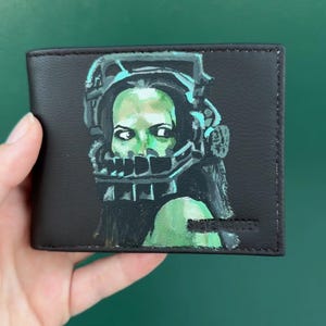 May include: A black bi-fold wallet featuring a hand-painted illustration of a person wearing a mechanical metal jaw trap device. The artwork uses shades of green and black to depict the face and the device. The Steve Madden logo is embossed.