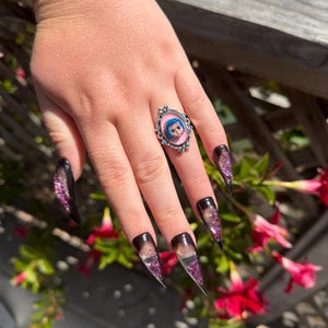 May include: A silver ring with a pink oval face featuring a cartoon character with blue hair and eyes. The ring is on a hand with long, pointed, black and purple glitter nails.
