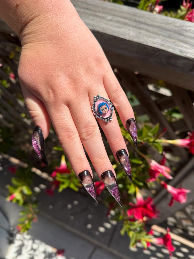 May include: A silver ring with a pink oval face featuring a cartoon character with blue hair and eyes. The ring is on a hand with long, pointed, black and purple glitter nails.