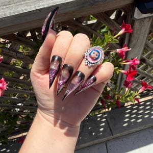 May include: A hand with long, pointed, black and purple glitter acrylic nails. A silver ring with a pink background and a cartoon face is on the ring finger. The background includes a wooden fence and red flowers.