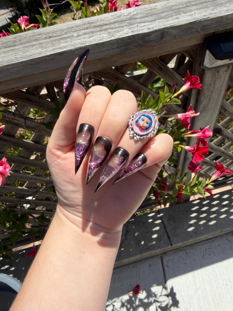 May include: A hand with long, pointed, black and purple glitter acrylic nails. A silver ring with a pink background and a cartoon face is on the ring finger. The background includes a wooden fence and red flowers.