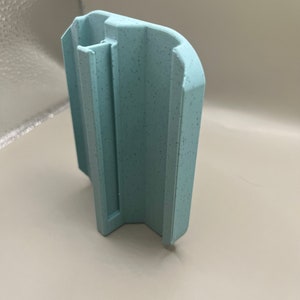 Puede incluir: Un objeto de pl&aacute;stico azul claro texturizado con una forma &uacute;nica. Tiene un cuerpo rectangular con una parte superior curvada y un canal vertical. La superficie presenta peque&ntilde;as motas oscuras.