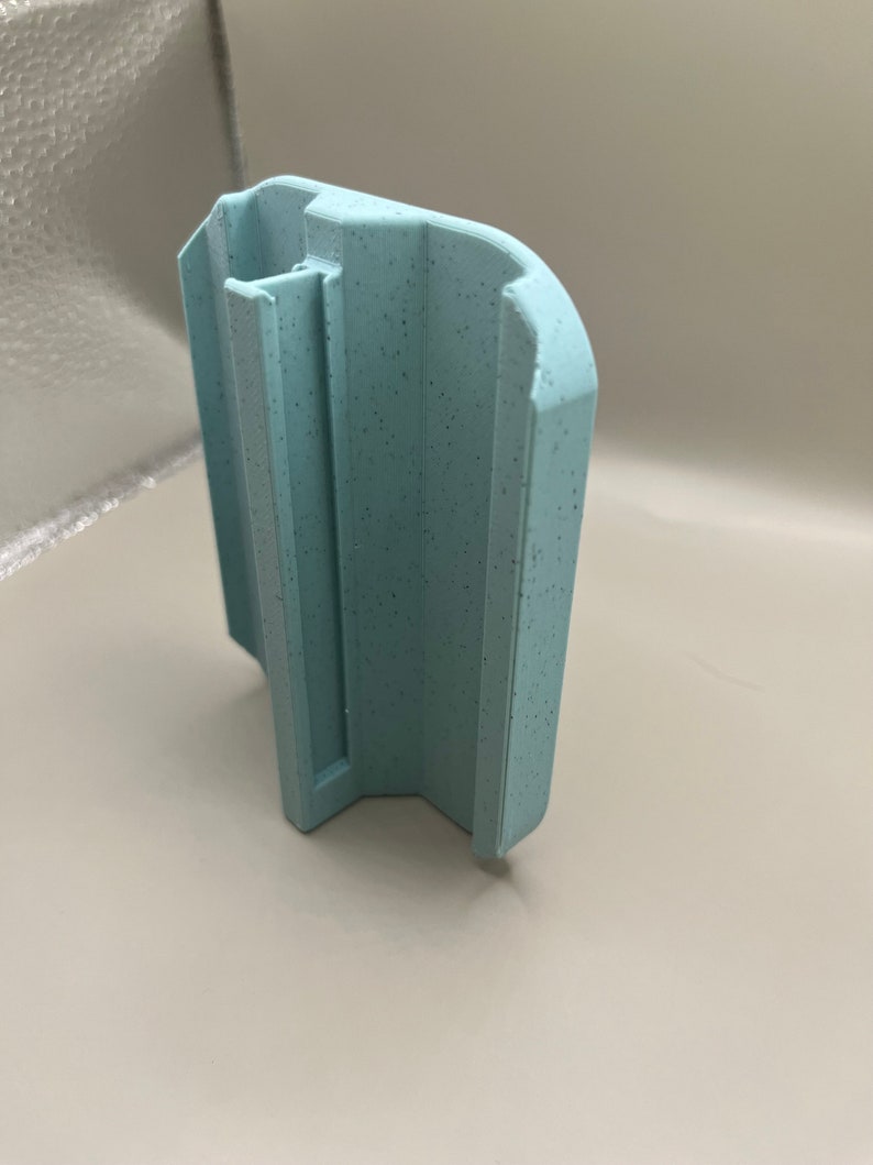 Puede incluir: Un objeto de pl&aacute;stico azul claro texturizado con una forma &uacute;nica. Tiene un cuerpo rectangular con una parte superior curvada y un canal vertical. La superficie presenta peque&ntilde;as motas oscuras.