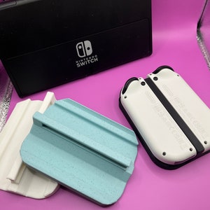 Puede incluir: Una consola Nintendo Switch negra y mandos Joy-Con blancos se muestran con dos soportes impresos en 3D, uno blanco y otro azul claro. Los soportes est&aacute;n dise&ntilde;ados para sujetar los mandos.