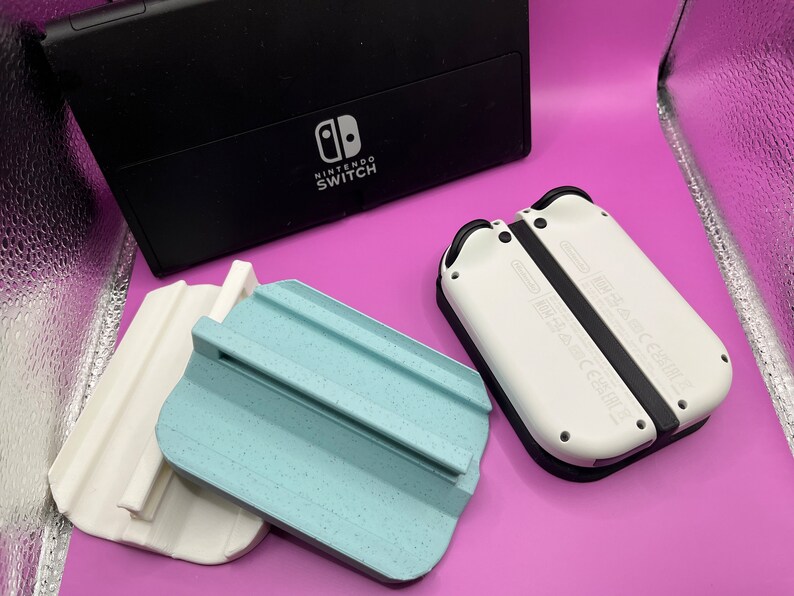 Puede incluir: Una consola Nintendo Switch negra y mandos Joy-Con blancos se muestran con dos soportes impresos en 3D, uno blanco y otro azul claro. Los soportes est&aacute;n dise&ntilde;ados para sujetar los mandos.
