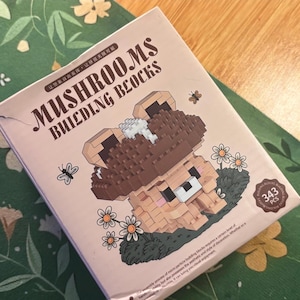May include: A box of "Mushrooms Building Blocks" featuring a mushroom-shaped bear illustration. The box is light pink with brown text and a cartoon bear with a mushroom cap. The box also has a small badge that says "343 pcs".