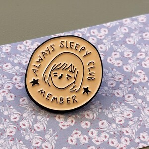 May include: A round, tan enamel pin with a black border. The pin features a cartoon illustration of a person with closed eyes and the text "ALWAYS SLEEPY CLUB MEMBER" in black. Two black stars flank the illustration.