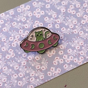 May include: A whimsical enamel pin featuring a green cat piloting a pink and green UFO. The pin has a silver outline and crescent moon details. The pin is set against a floral patterned background.