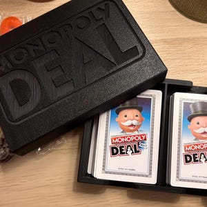 May include: A black Monopoly Deal card game box with the game title embossed on the lid. The box is open, revealing a stack of cards with the Monopoly Deal logo and a cartoon character. Wrapped candies are scattered around the box.