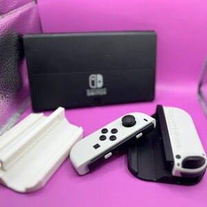 Puede incluir: Una consola Nintendo Switch en blanco y negro con mandos Joy-Con blancos y una base de carga. La consola est&aacute; sobre un fondo morado, con un soporte blanco y una superficie reflectante plateada.