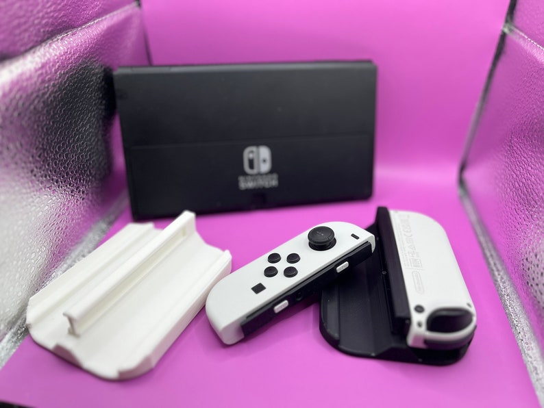 Puede incluir: Una consola Nintendo Switch en blanco y negro con mandos Joy-Con blancos y una base de carga. La consola est&aacute; sobre un fondo morado, con un soporte blanco y una superficie reflectante plateada.