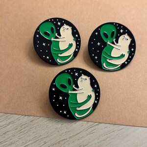 May include: Three round enamel pins featuring a white cat embracing a green alien against a black starry background. The pins are on a brown surface with a light wood grain pattern.