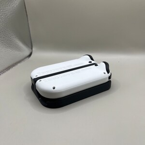 Puede incluir: Dos mandos de juego blancos y negros apilados. Los mandos tienen forma redondeada con detalles negros y peque&ntilde;os agujeros. El mando superior es blanco y el inferior es negro. Probablemente para una consola de juegos.