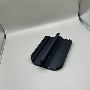 Puede incluir: Un soporte negro para tel&eacute;fono, de forma rectangular, con un soporte central elevado y lados inclinados. Dise&ntilde;ado para sostener un tel&eacute;fono en posici&oacute;n vertical, ofrece un &aacute;ngulo de visi&oacute;n conveniente. El soporte est&aacute; hecho de un material negro mate.