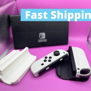 Puede incluir: Una base negra de Nintendo Switch y mandos Joy-Con blancos se muestran sobre una superficie rosa. Los mandos tienen botones negros y un joystick negro. El texto "Fast Shipping" est&aacute; en un rect&aacute;ngulo azul.