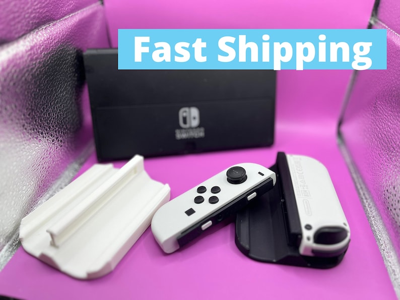 Puede incluir: Una base negra de Nintendo Switch y mandos Joy-Con blancos se muestran sobre una superficie rosa. Los mandos tienen botones negros y un joystick negro. El texto "Fast Shipping" est&aacute; en un rect&aacute;ngulo azul.