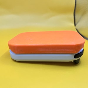 Puede incluir: Un objeto rectangular de dos tonos con una parte superior naranja y una base blanca. El objeto tiene esquinas redondeadas y un clip negro en un lado. El fondo es amarillo liso.