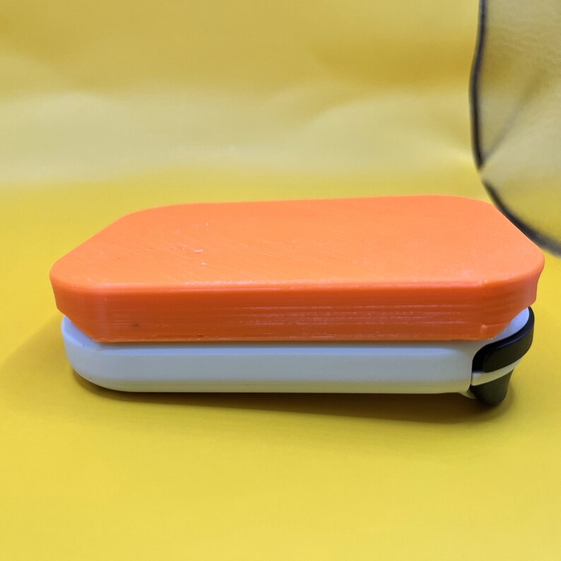 Puede incluir: Un objeto rectangular de dos tonos con una parte superior naranja y una base blanca. El objeto tiene esquinas redondeadas y un clip negro en un lado. El fondo es amarillo liso.