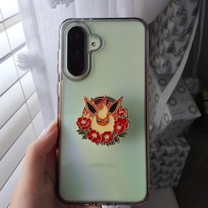 Puede incluir: Un smartphone Samsung de color verde claro en una funda protectora transparente con un soporte circular de esmalte. El soporte muestra una criatura estilizada de color naranja y crema con orejas grandes rodeada de flores rojas.