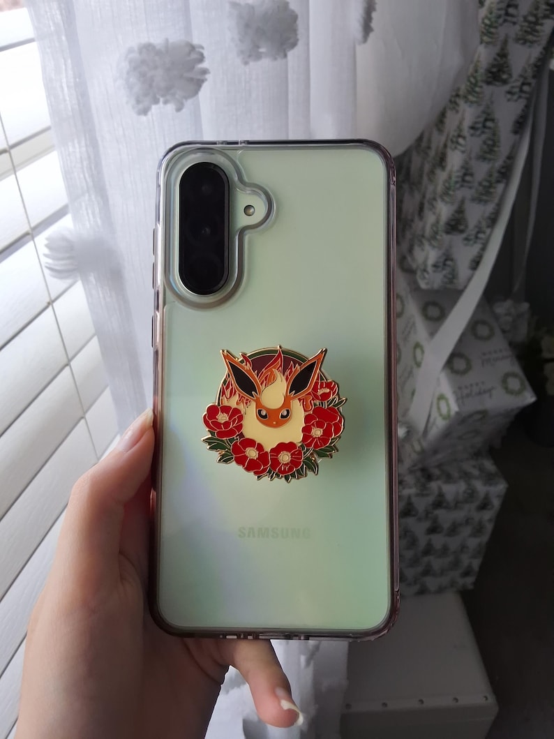 Puede incluir: Un smartphone Samsung de color verde claro en una funda protectora transparente con un soporte circular de esmalte. El soporte muestra una criatura estilizada de color naranja y crema con orejas grandes rodeada de flores rojas.