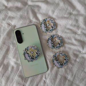 May include: A light green smartphone with a circular phone grip attached, featuring a design of a white whale swimming through blue and white waves. Three additional identical circular phone grips are arranged to the right of the device.