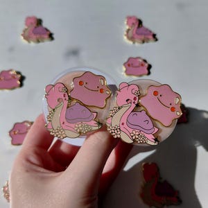 Puede incluir: Broches de esmalte rosa y dorado con un diseño inspirado en Ditto y Pokémon. Los broches tienen un acabado brillante y detalles florales. Varios broches están dispersos sobre una superficie blanca. Los broches miden aproximadamente 2,5 cm.