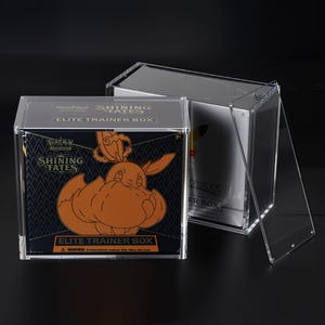 May include: Clear acrylic display case for a "Shining Fates Elite Trainer Box". The box features a black background with orange artwork of a Pokemon character. The text "Shining Fates" and "Elite Trainer Box" are visible on the box.