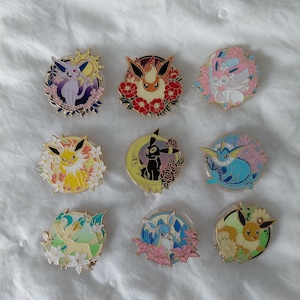May include: Nine colorful enamel pins featuring various Pokémon characters surrounded by floral designs. The pins showcase a variety of colors, including pink, blue, yellow, and green, with intricate details and a glossy finish. The pins are round and have a metal border.