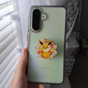 Puede incluir: Una funda de smartphone verde claro con un soporte circular de esmalte. El soporte muestra una criatura amarilla de pelaje puntiagudo y orejas grandes, rodeada de flores blancas sobre un fondo naranja brillante con bordes dorados.