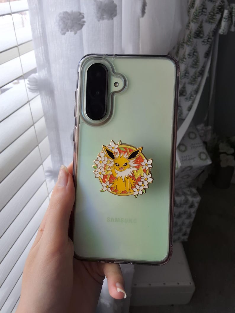 Puede incluir: Una funda de smartphone verde claro con un soporte circular de esmalte. El soporte muestra una criatura amarilla de pelaje puntiagudo y orejas grandes, rodeada de flores blancas sobre un fondo naranja brillante con bordes dorados.