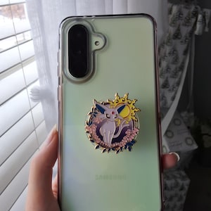 Puede incluir: Una funda transparente para tel&eacute;fono Samsung con un accesorio circular de esmalte en la parte posterior. El accesorio muestra una criatura de color morado claro con orejas grandes, una gema roja en la frente y un sol amarillo, rodeado de flores.