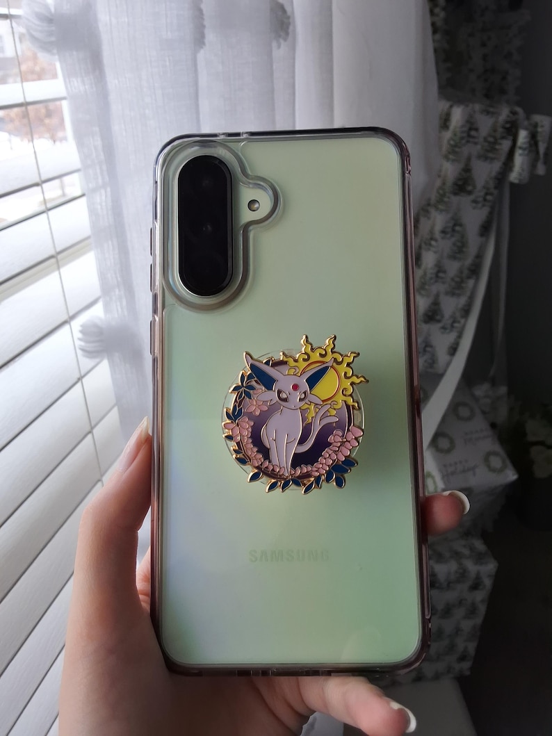 Puede incluir: Una funda transparente para tel&eacute;fono Samsung con un accesorio circular de esmalte en la parte posterior. El accesorio muestra una criatura de color morado claro con orejas grandes, una gema roja en la frente y un sol amarillo, rodeado de flores.