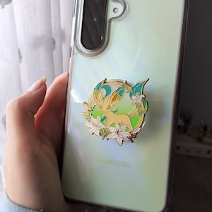 Puede incluir: Una funda transparente para tel&eacute;fono Samsung con acabado iridiscente que presenta un pin de esmalte con borde dorado de una criatura amarilla con detalles en verde azulado rodeada de flores de lirio blancas.