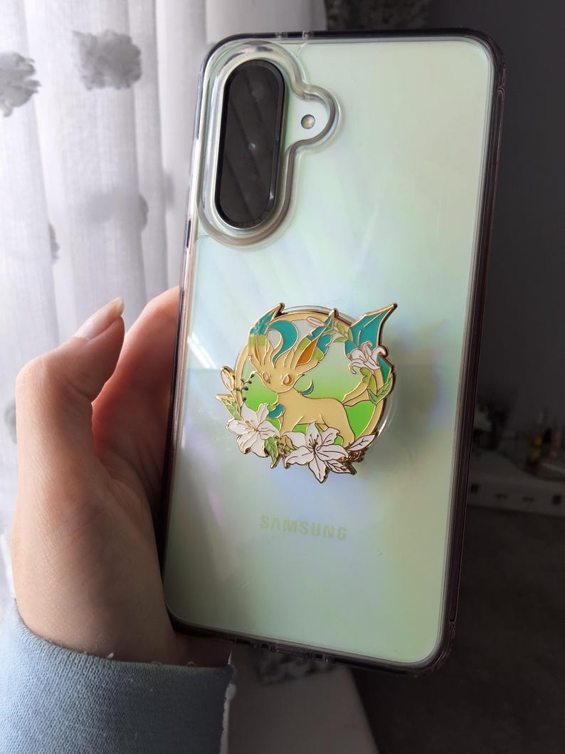 Puede incluir: Una funda transparente para tel&eacute;fono Samsung con acabado iridiscente que presenta un pin de esmalte con borde dorado de una criatura amarilla con detalles en verde azulado rodeada de flores de lirio blancas.